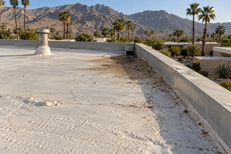 Illustrative view along the edge of a white-coated flat roof, sand and grit drifted against the edge metal at a scupper, a vent stack and mountains beyond