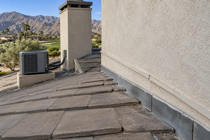 Illustrative close view of concrete roof tile meeting a tall stucco wall at its flashing, a chimney and an air-conditioning unit on a platform, a fairway and mountains beyond