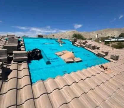 A tile roof part-way through a reroof: blue Tyvek underlayment laid across the deck, tiles stacked on the left and beside it, hills under a clear blue sky