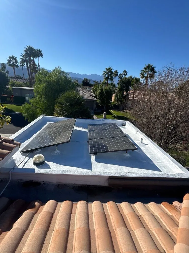 After: a flat roof finished in clean white spray foam, with two solar panels, palms and mountains beyond