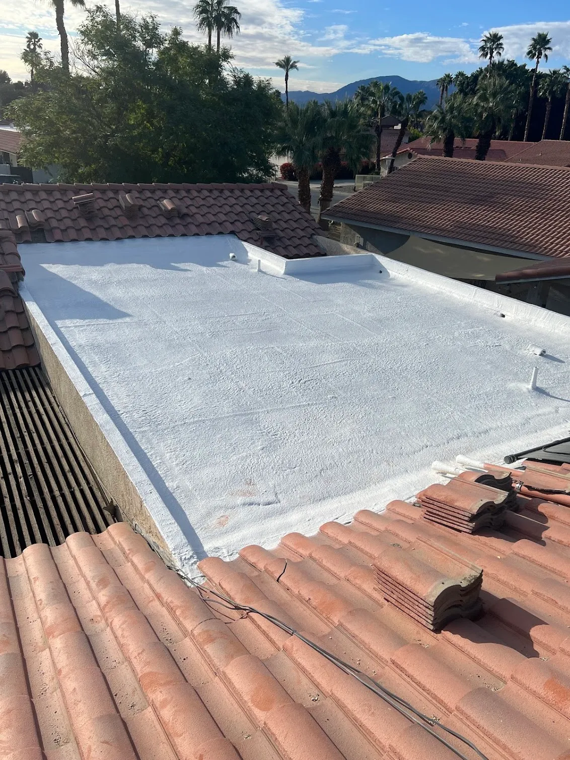A flat roof finished in bright white spray foam, framed on every side by terracotta barrel tile, with palms and the desert mountains behind