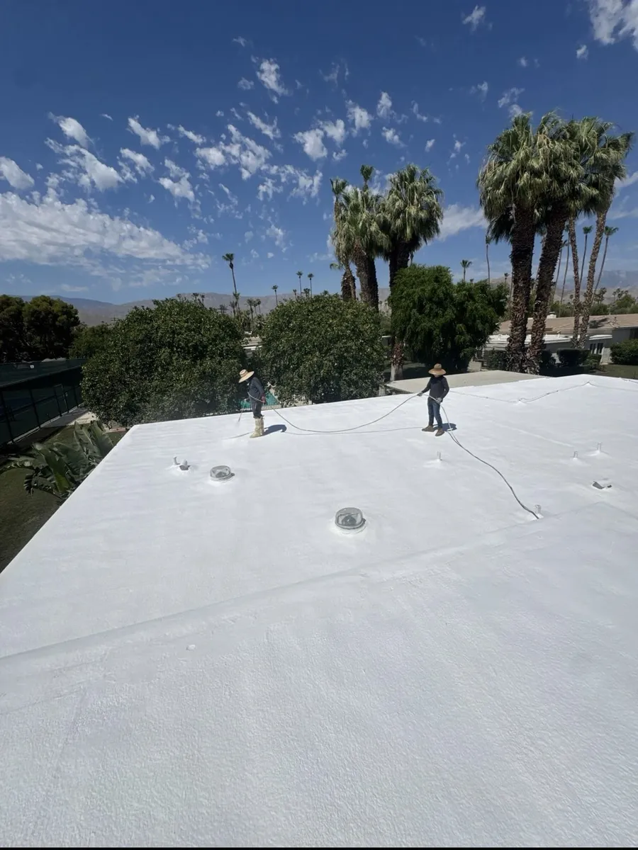 Two roofers finishing a freshly coated white spray foam roof on a valley home, palms and mountains behind