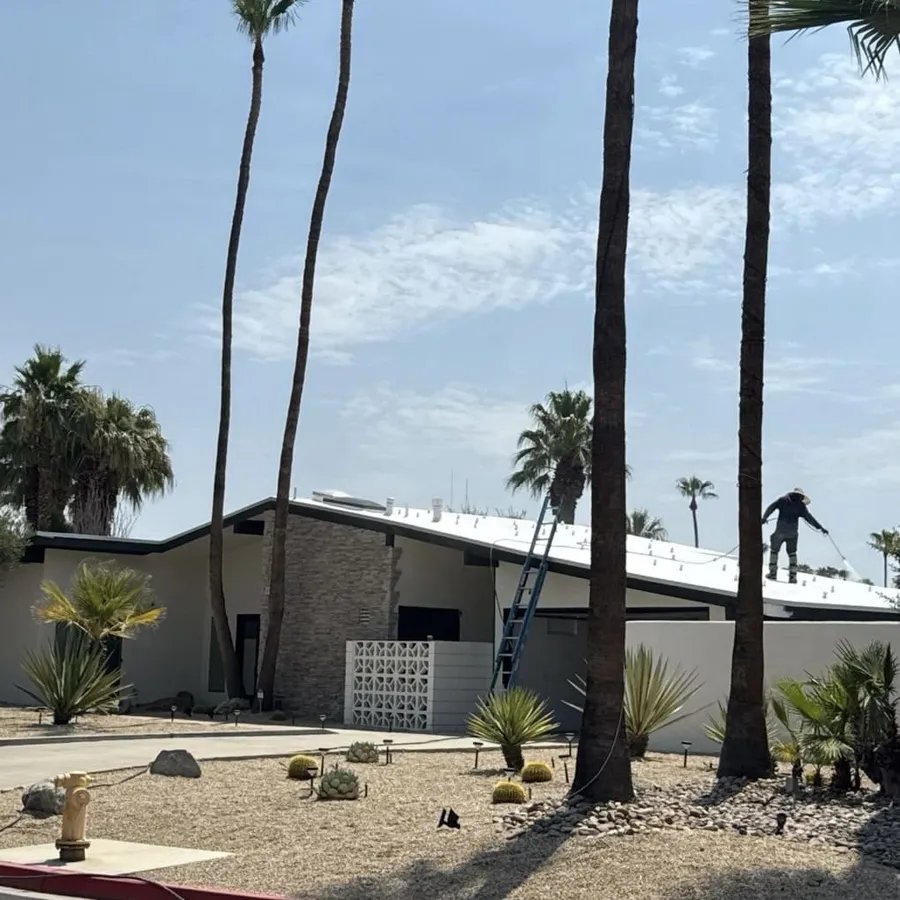 A mid-century desert home from the street with a roofer working on its white spray foam roof, ladder at the eave