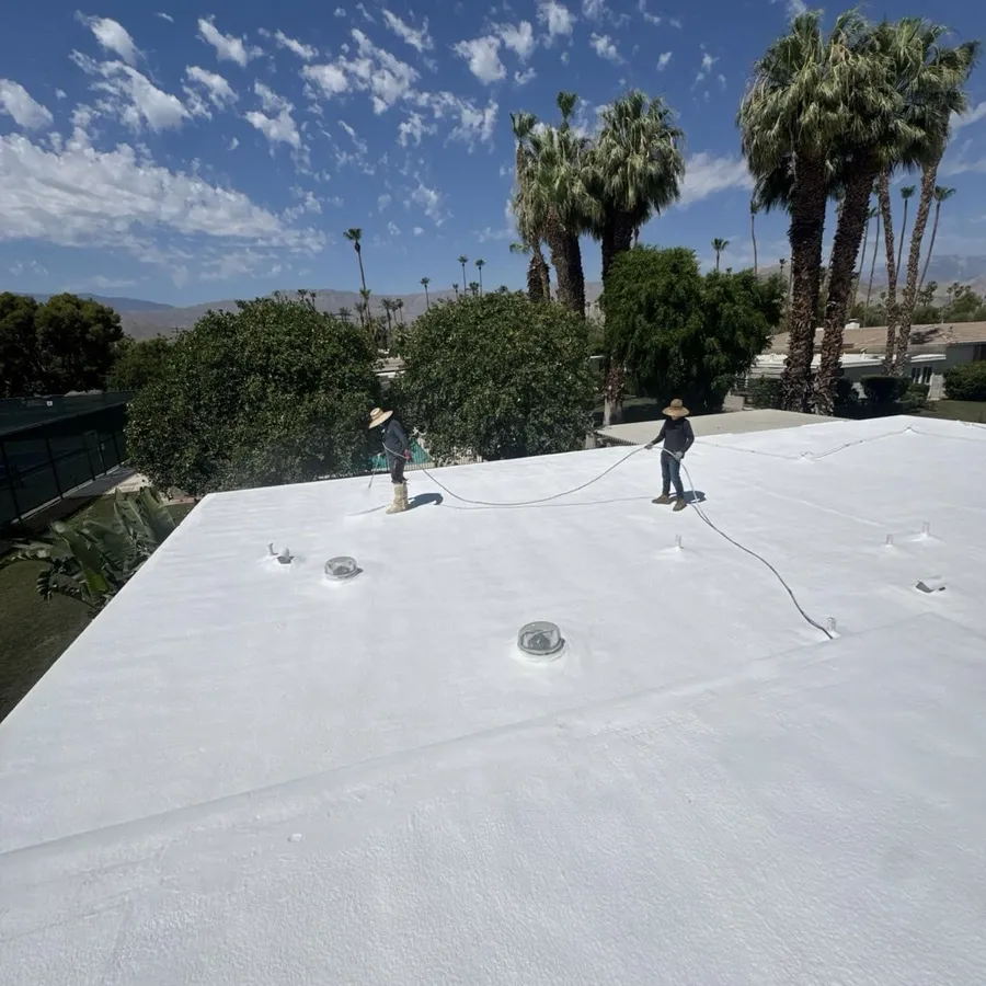 Two roofers working across a newly coated white spray foam roof, palms and greenery behind