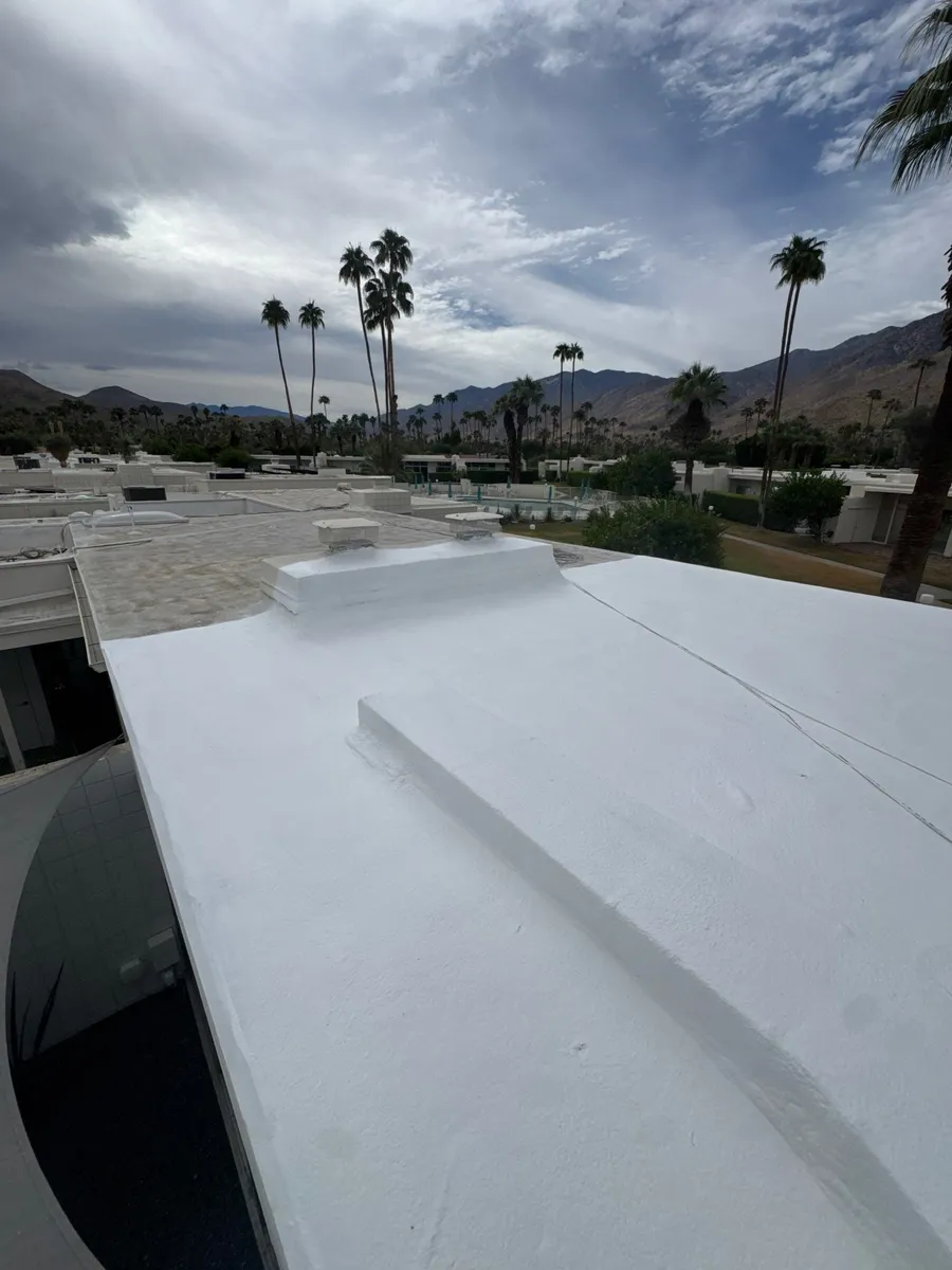 A flat roof part-way through renewal: fresh white spray foam coating beside the weathered grey surface still to be done, palms and mountains under cloud