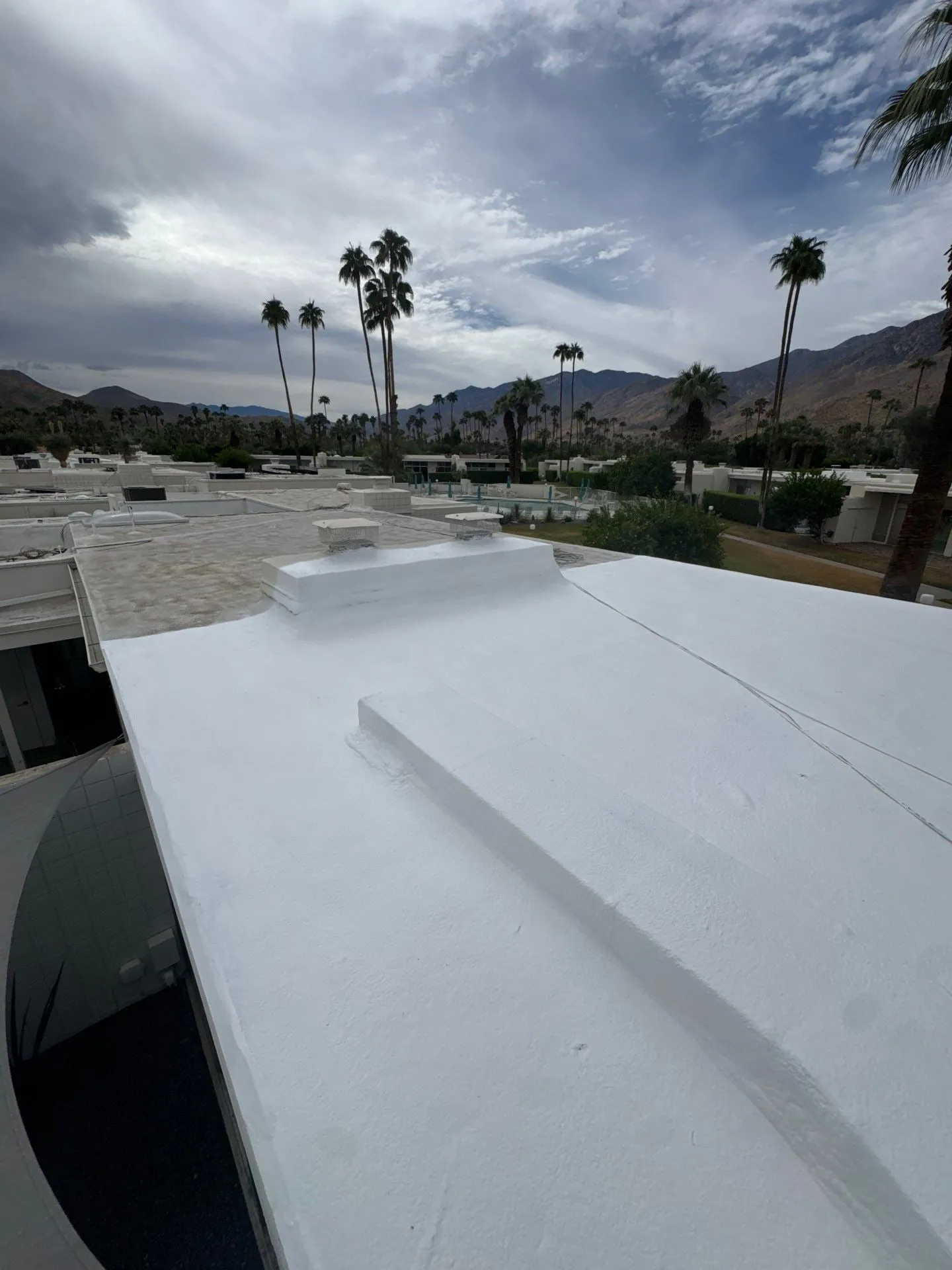 A flat roof part-way through renewal: fresh white SPF coating beside the weathered grey surface still to be done, palms and mountains under cloud