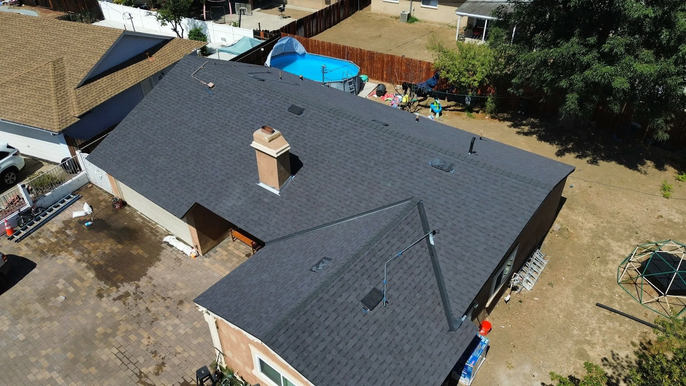 Drone view of a single-storey home with a new charcoal shingle roof, chimney flashed and vents in place, a backyard pool beyond