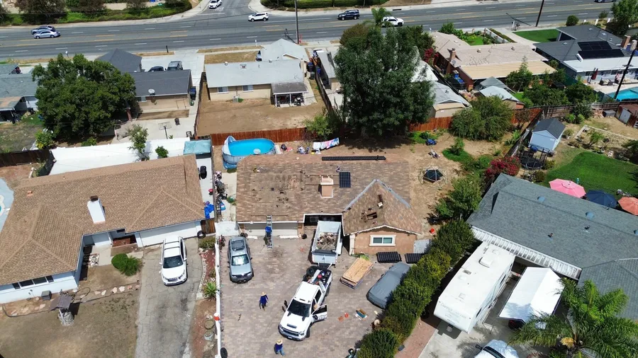 Drone view of a shingle tear-off under way: old brown shingles partly stripped, a debris trailer in the driveway and the crew at work