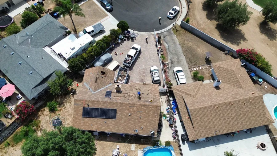 Overhead drone view of a home at the start of a reroof, its worn brown shingles and solar panels still in place, a debris trailer in the driveway